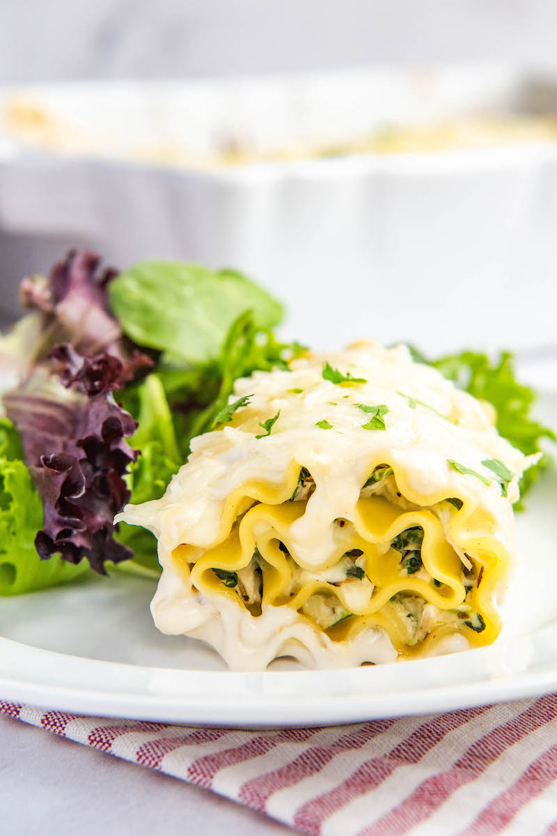 A white chicken lasagna rollup sits on a plate next to a leafy salad