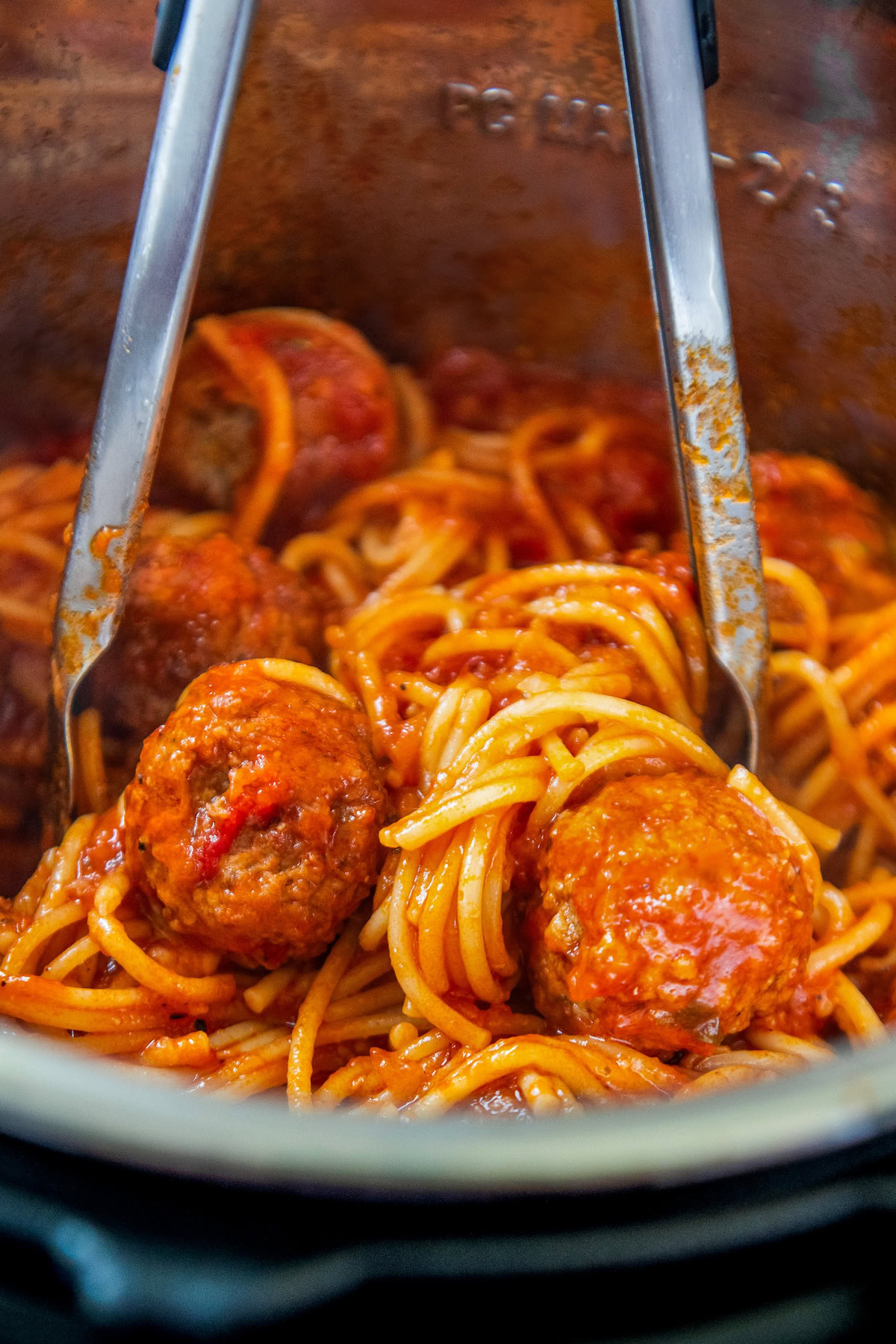 Tongs are tossing marinara sauce, spaghetti, and meatballs together in a pressure cooker.