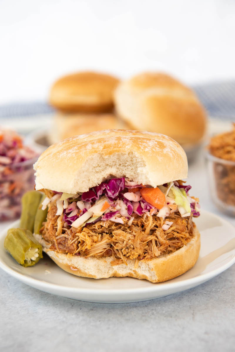 A pulled pork sandwich sits on a white plate next to pickled okra