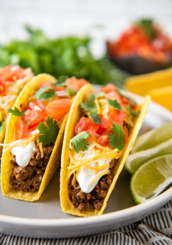 Hard shell tacos are on a white plate with sliced limes