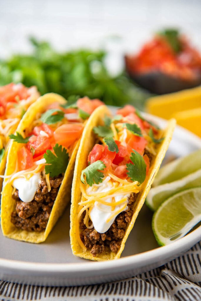 Hard shell tacos are on a white plate with sliced limes