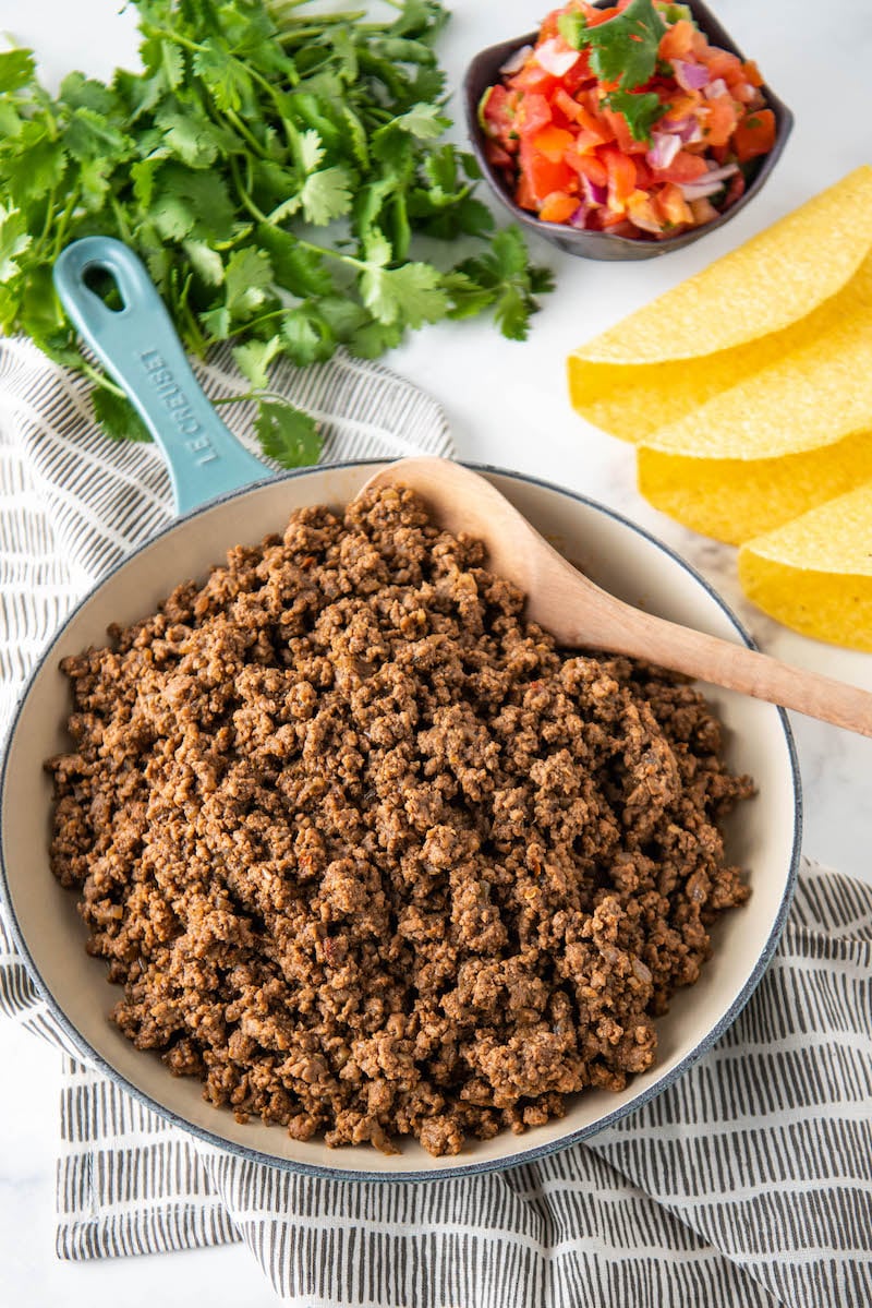 A skillet is filled with ground beef and is being stirred with a wooden spoon