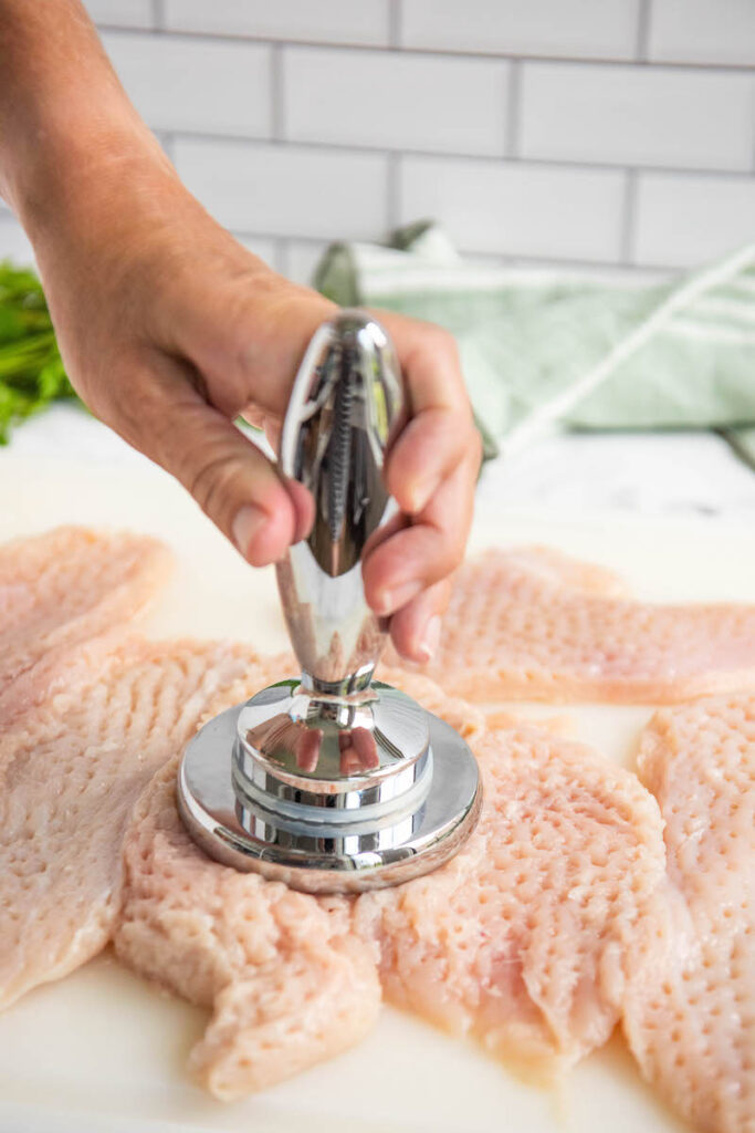 Chicken breasts on a cutting board being pounded thin with a meat tenderizer.