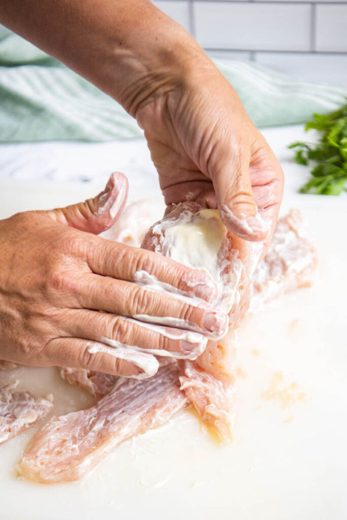 Thin chicken breasts on a cutting board being coated in mayo.