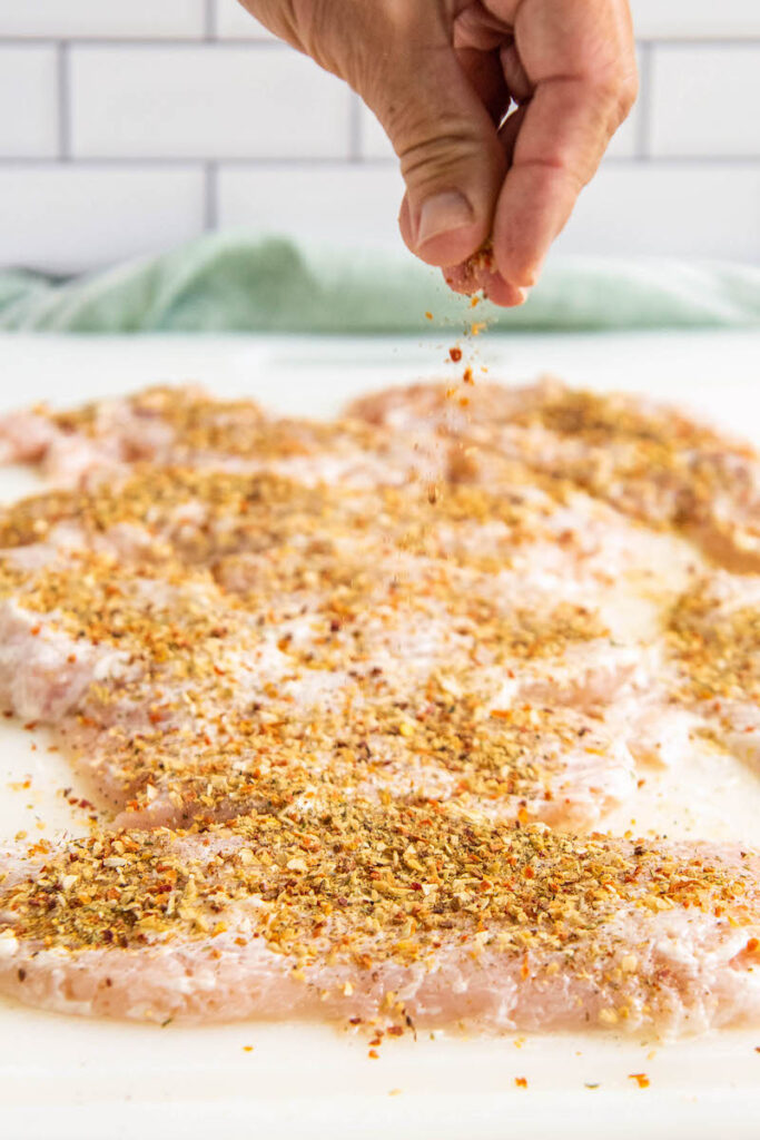 Chicken breasts on a cutting board being sprinkled with seasonings.