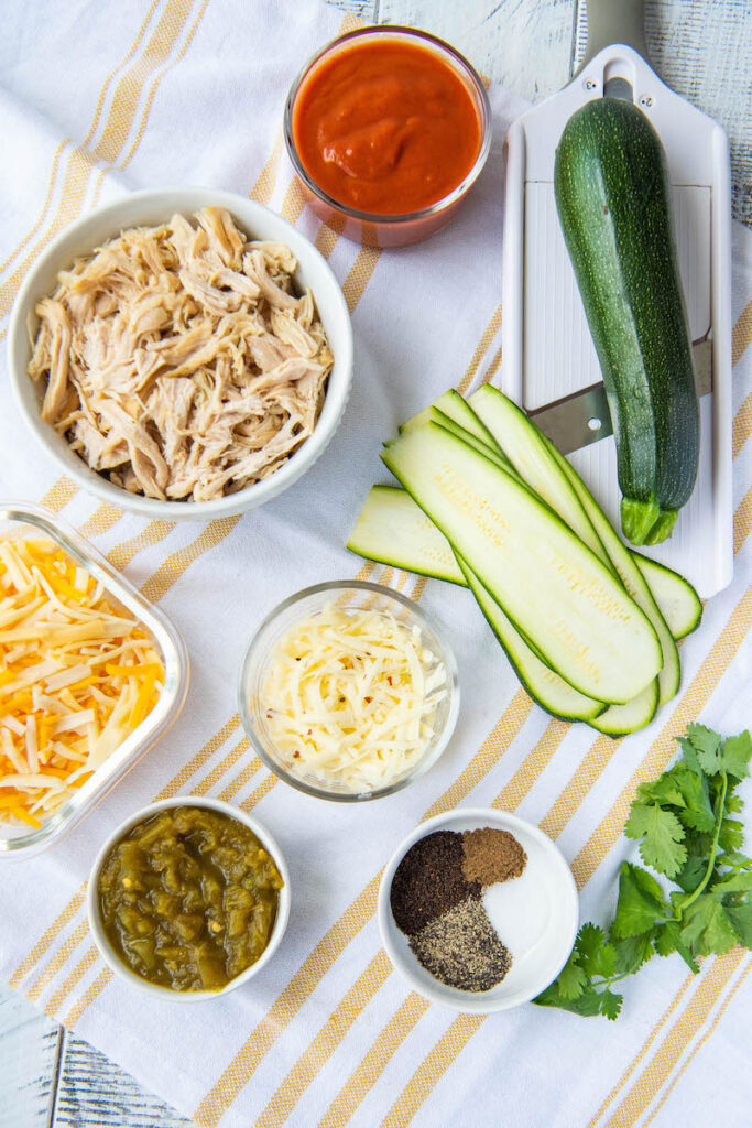 Chicken in a bowl. Cut zucchini. Cheese, seasoning, cilantro on a yellow and white tea towel.