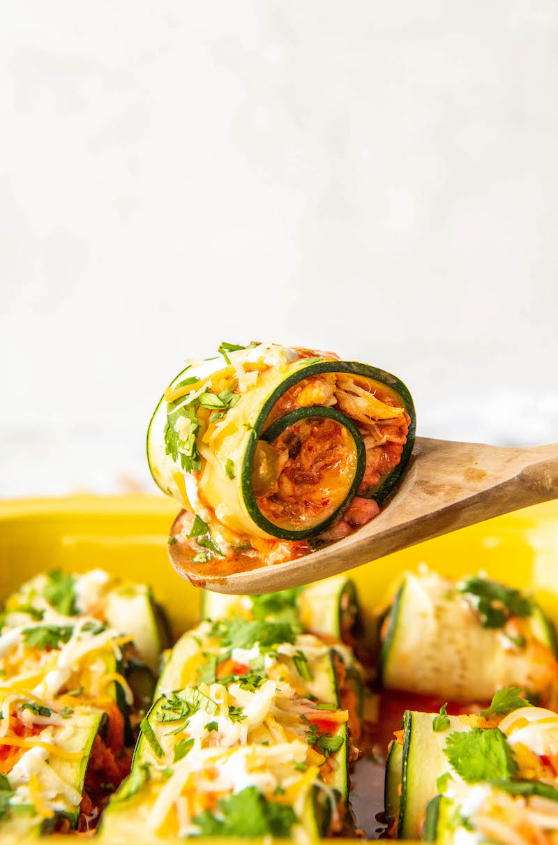 Zucchini enchiladas in a yellow baking dish and one on a wooden spoon.