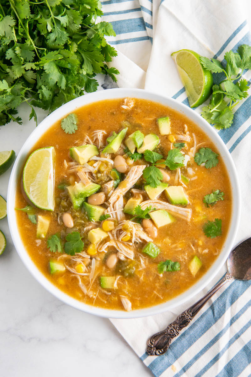 Overhead view of salsa verde chicken soup on a white and blue stripe napkin with lime and cilantro.