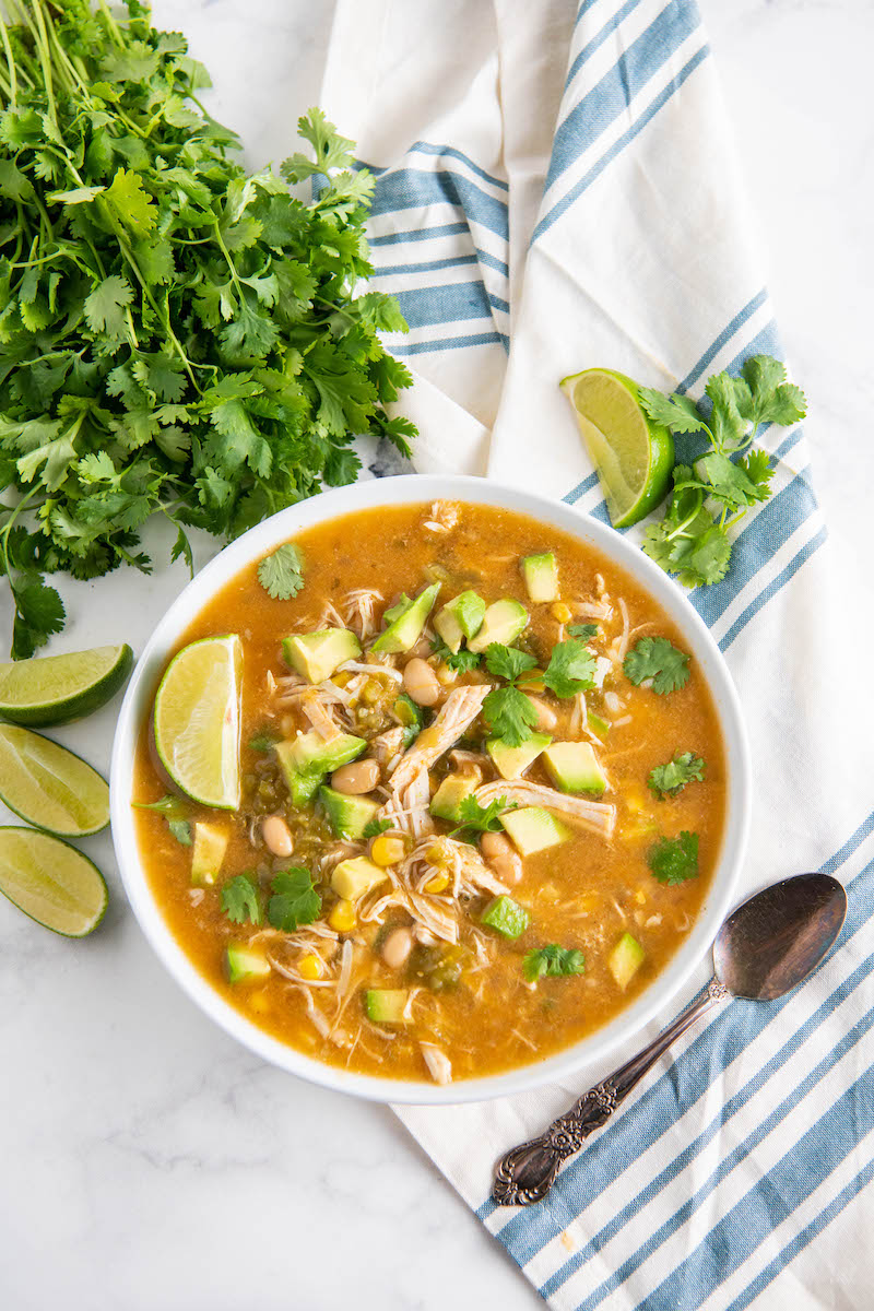 Overhead image of chicken crockpot soup with a napkin, spoon, cilantro, avocado and lime.