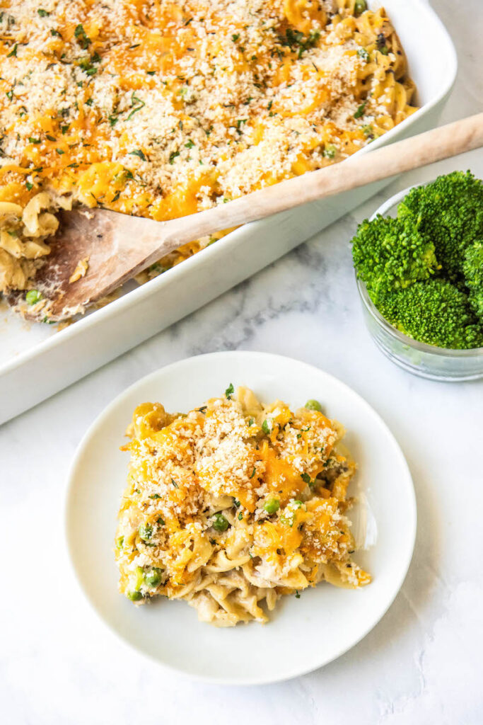 A plate with tuna noodle casserole is on a white tabletop, next to a pan full of the casserole.