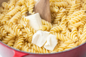 Up close image of cooked noodles in a red pot with cream cheese and a wooden spoon.