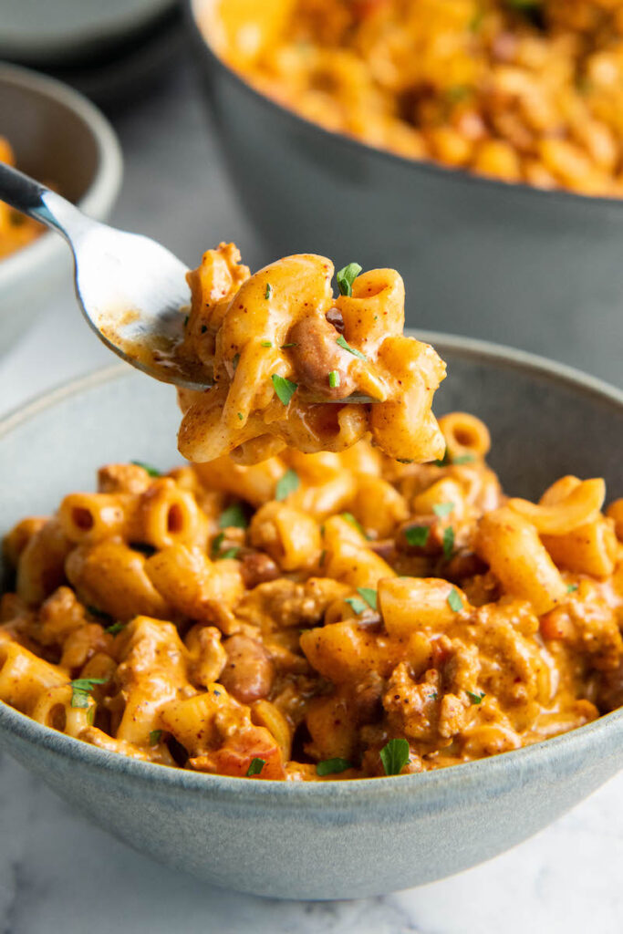 A fork is holding up a bite sized portion of chili Mac above a bowl.