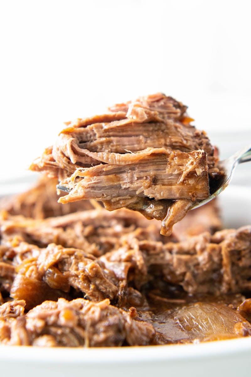 A serving spoon is holding perfectly cooked, flaky shredded beef above a crock pot.