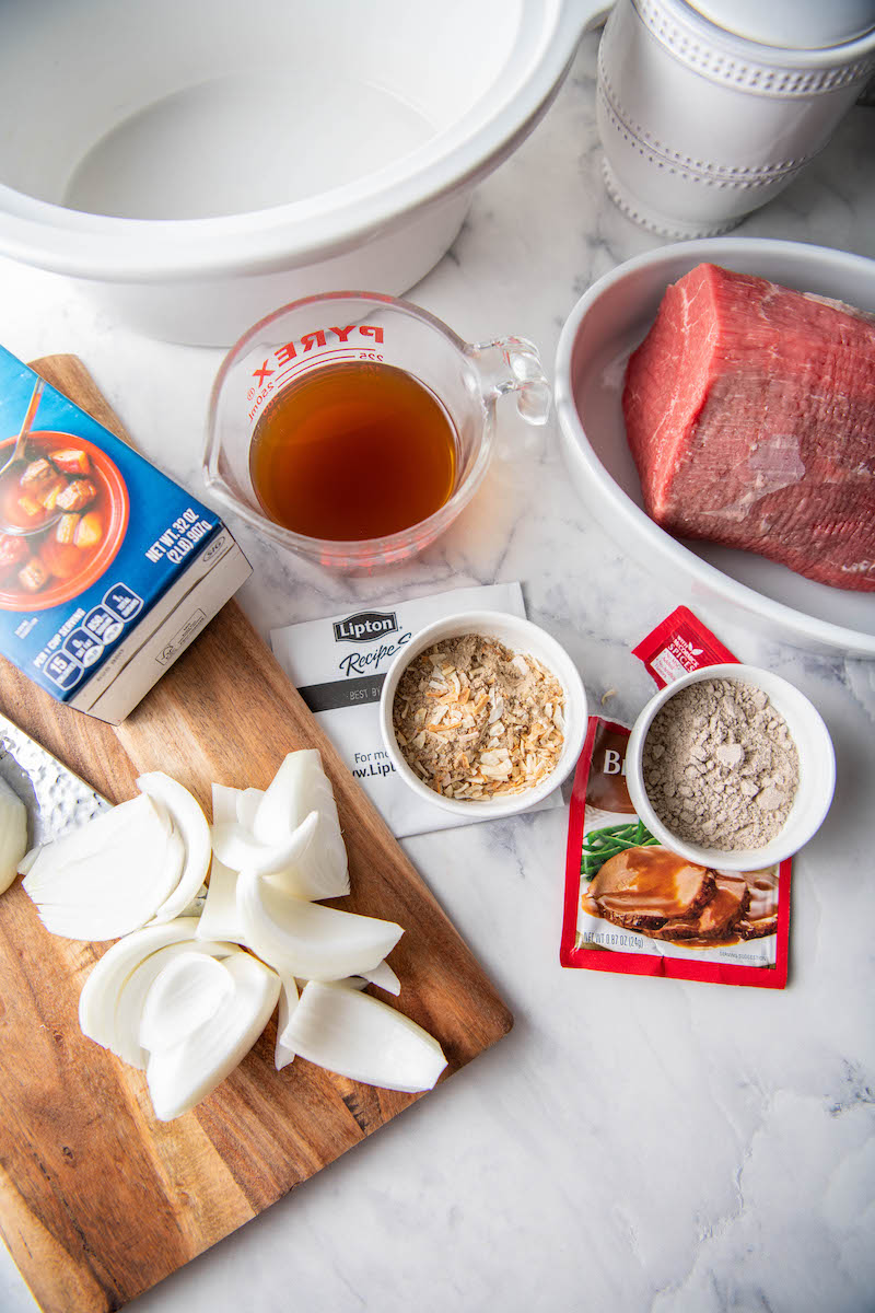 The ingredients for crockpot shredded beef are spread out on a white countertop.