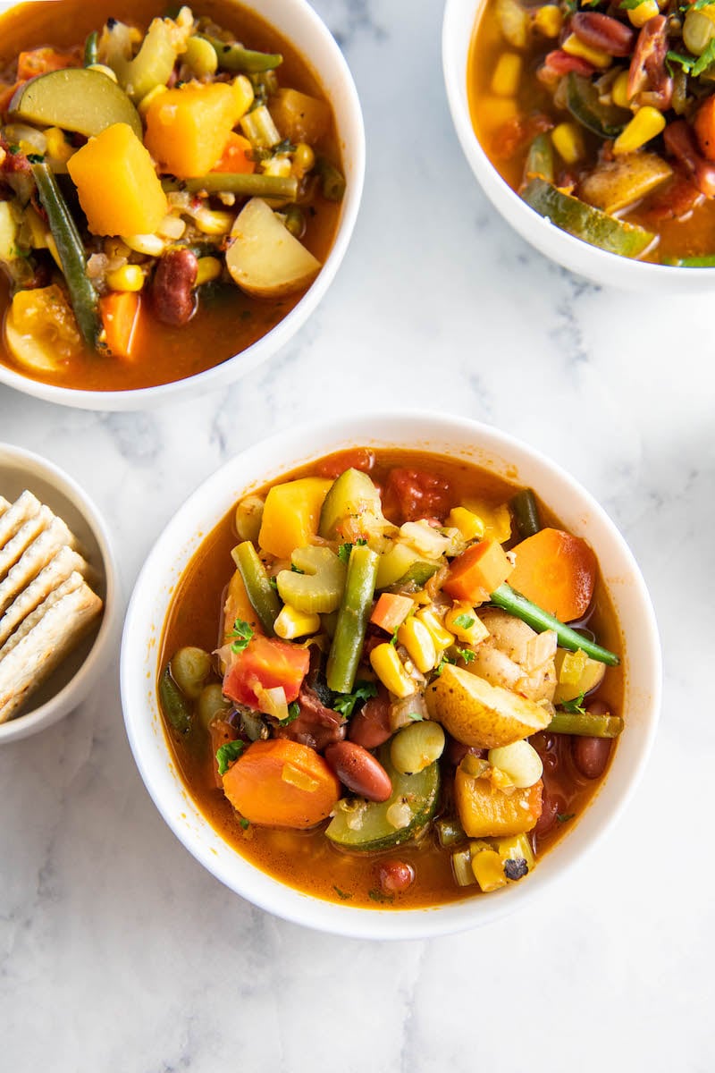 Three white bowls containing servings of vegetable soup are placed on a white surface.