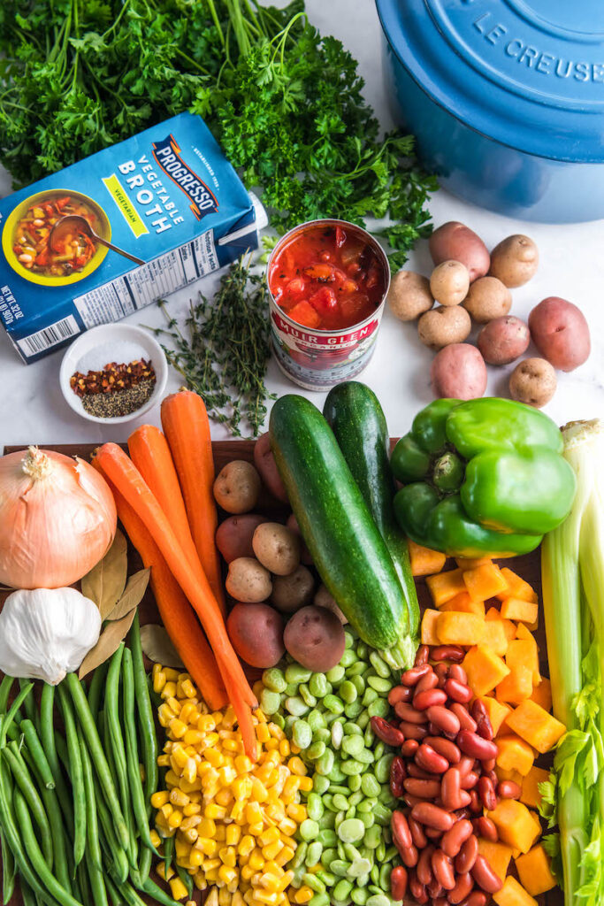 The ingredients for vegetable soup are spread out on a white surface.