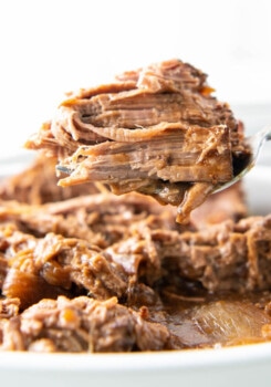 A serving spoon is holding perfectly cooked, flaky shredded beef above a crock pot.