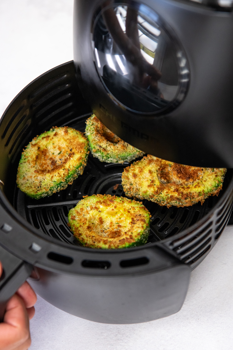 The air fryer basket is being opened, showing four cooked avocados.