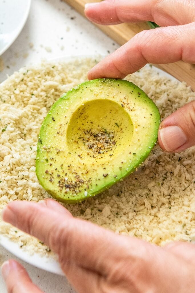 An avocado half is being coated in seasoned breadcrumbs.