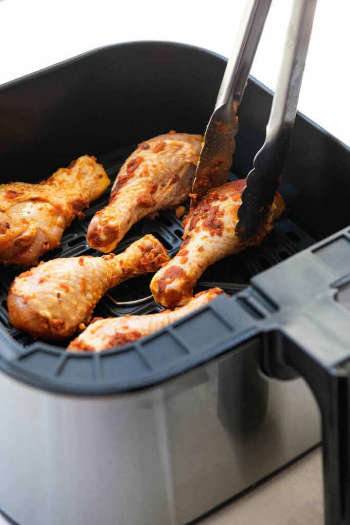 Chicken legs being placed into an air fryer basket with tongs.