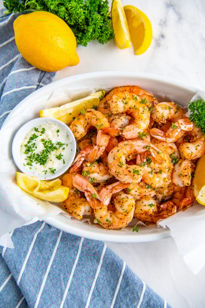 A white oval bowl contains a large serving of cooked shrimp and lemon wedges.