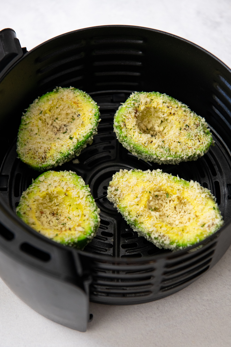 Four avocados with uncooked breadcrumbs are plated in the black air fryer basket.