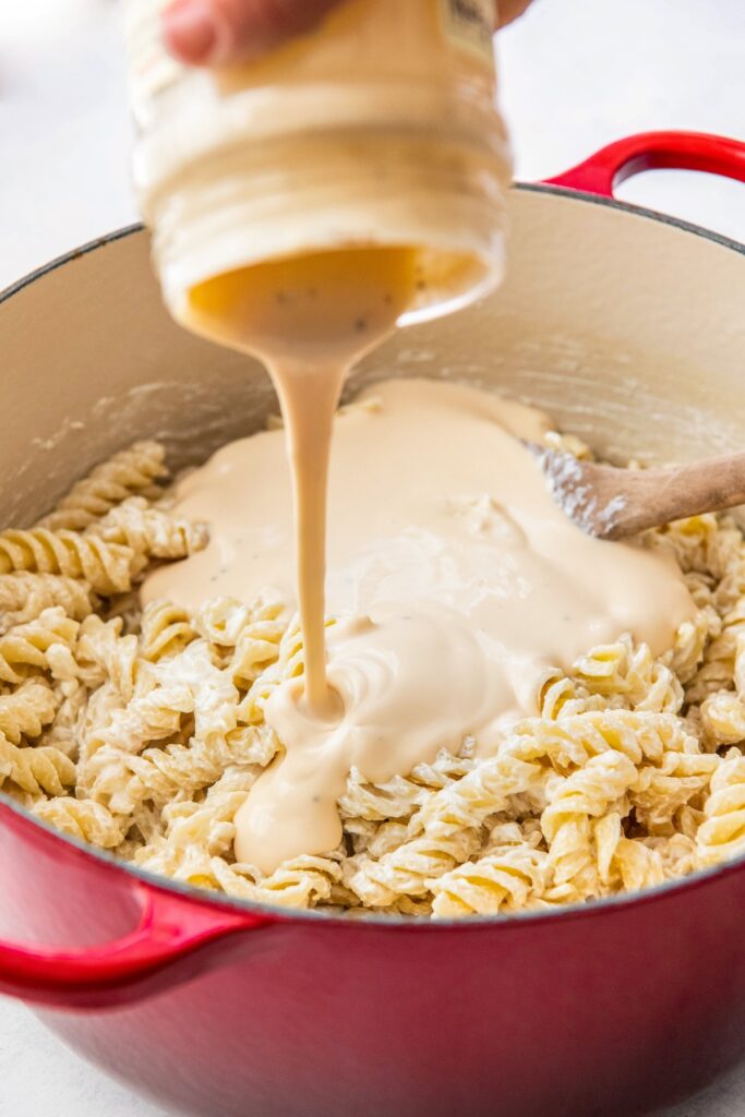 Alfredo sauce is being poured into a pot filled with cooked pasta.