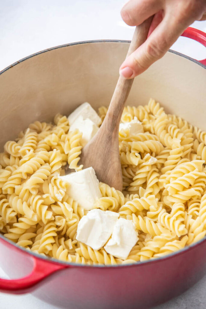 Cream cheese cubes are being mixed into a pot filled with cooked noodles.