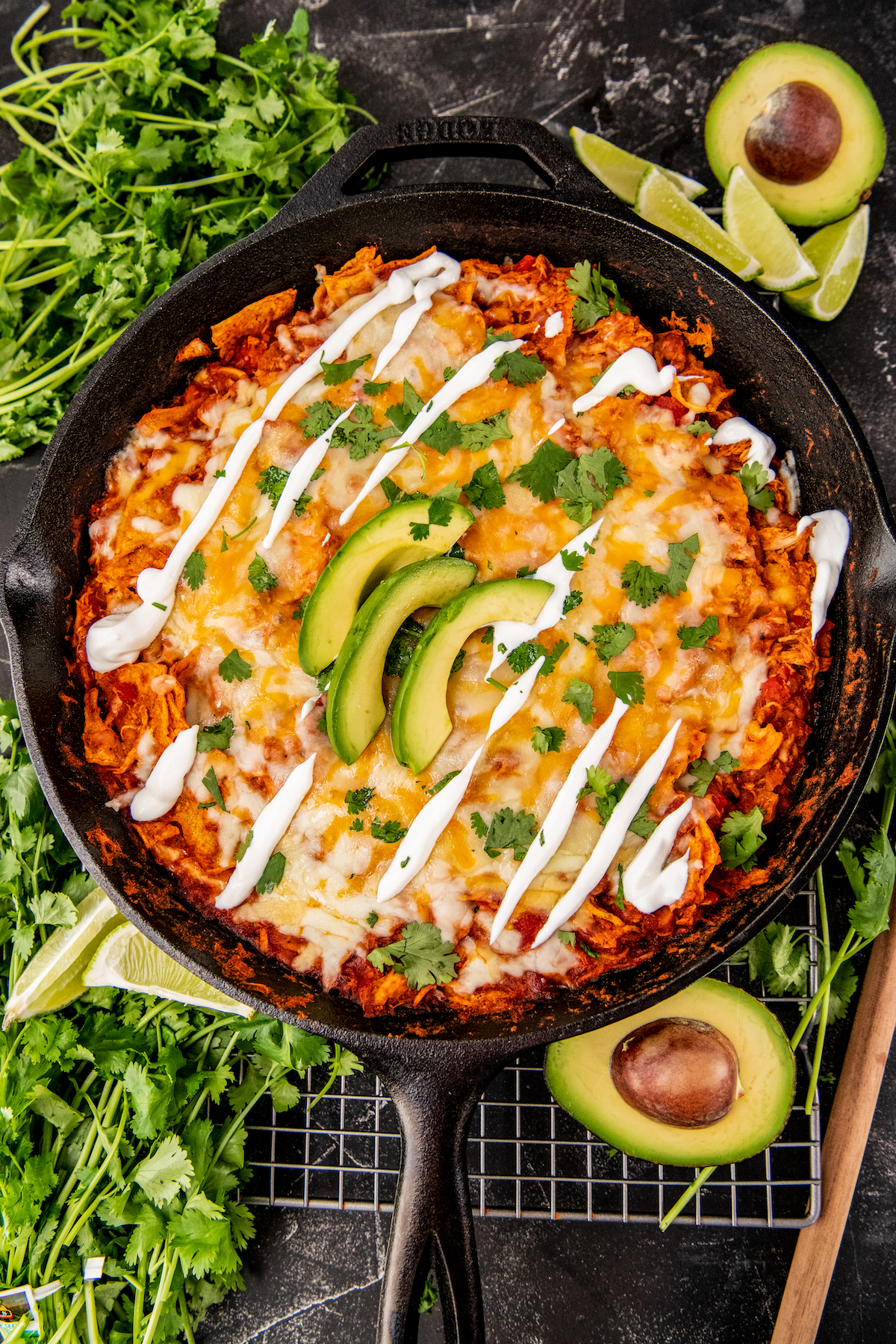 Avocado halves and bunches of cilantro are placed around a skillet full of enchiladas.