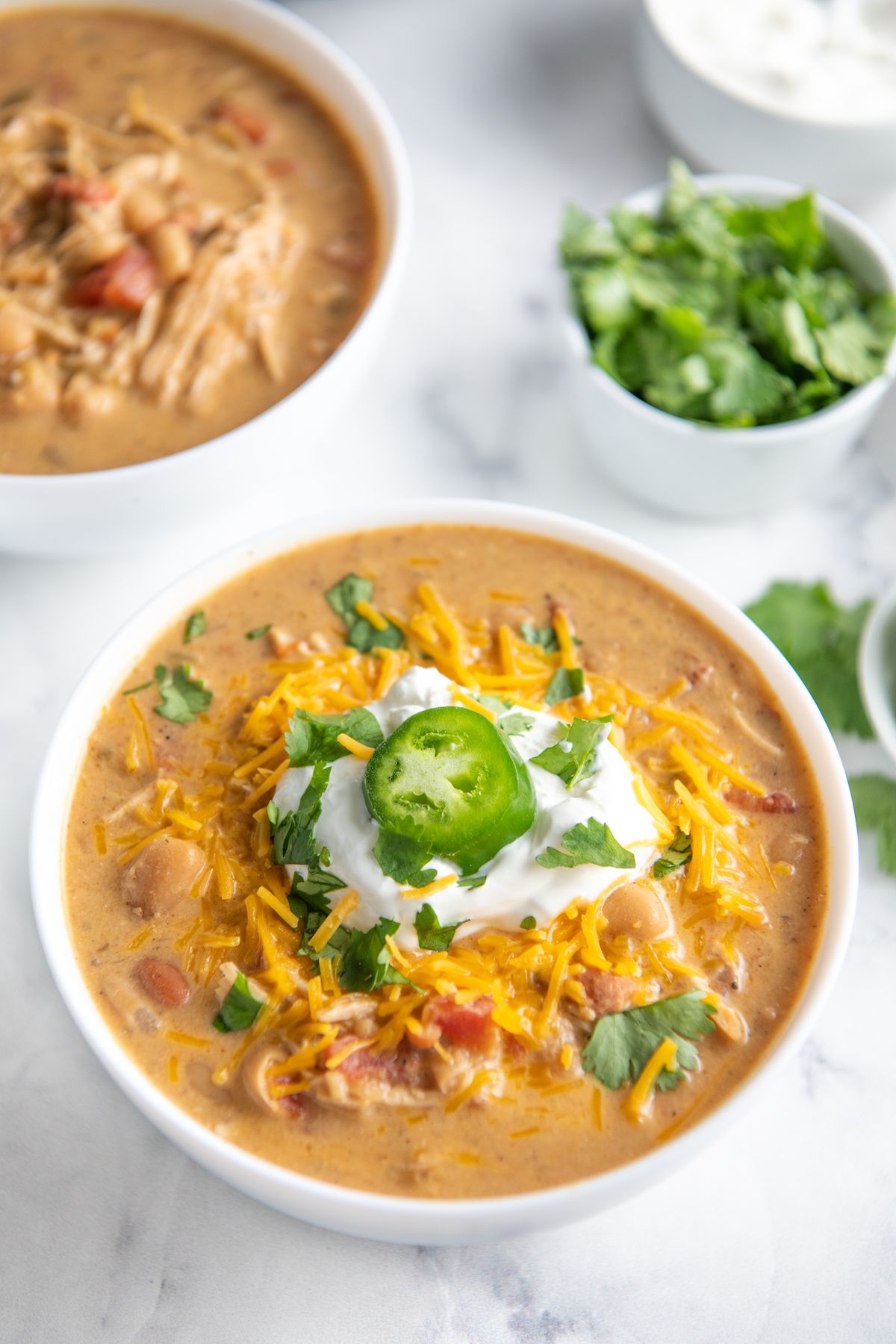 A white bowl with chicken chili topped with sour cream and jalapeno slice.