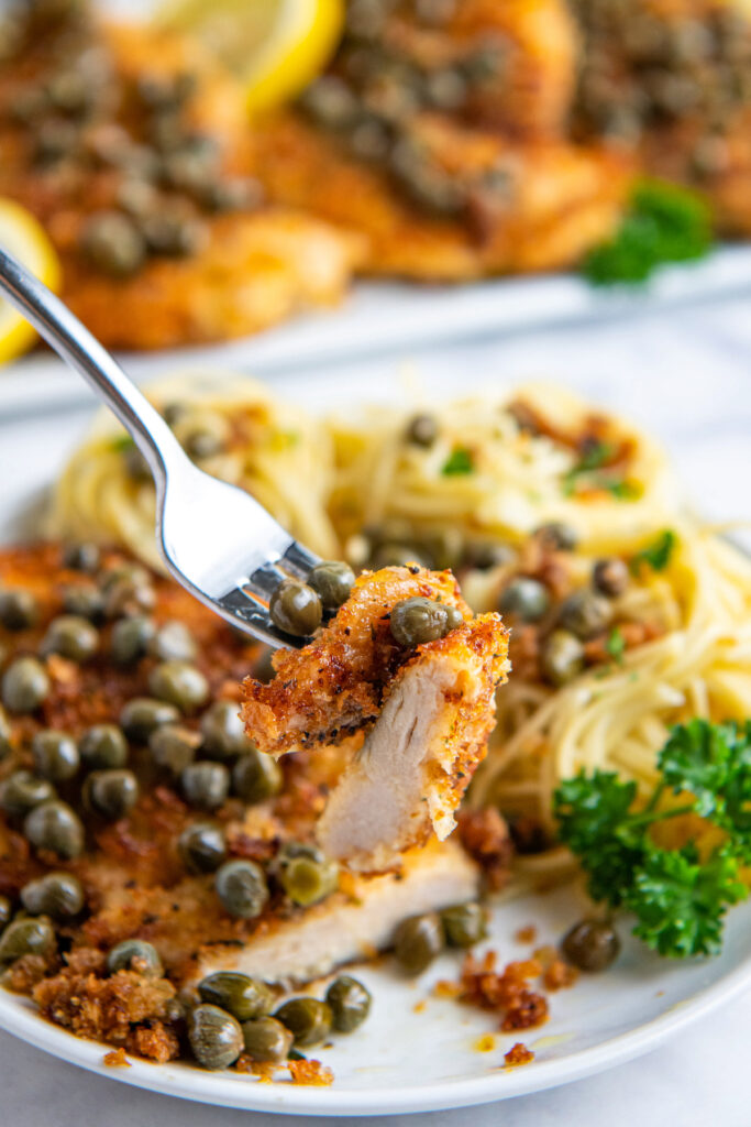 Chicken piccata on a fork with capers served from a plate with more chicken and pasta