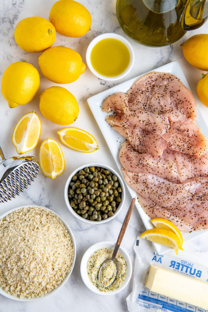 Lemons, capers, breadcrumbs, garlic salt and raw chicken breast on a plate