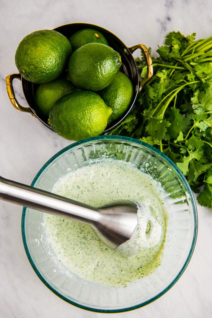 Cilantro lime crema in a glass bowl with an immersion blender. Cilantro and lime on the side