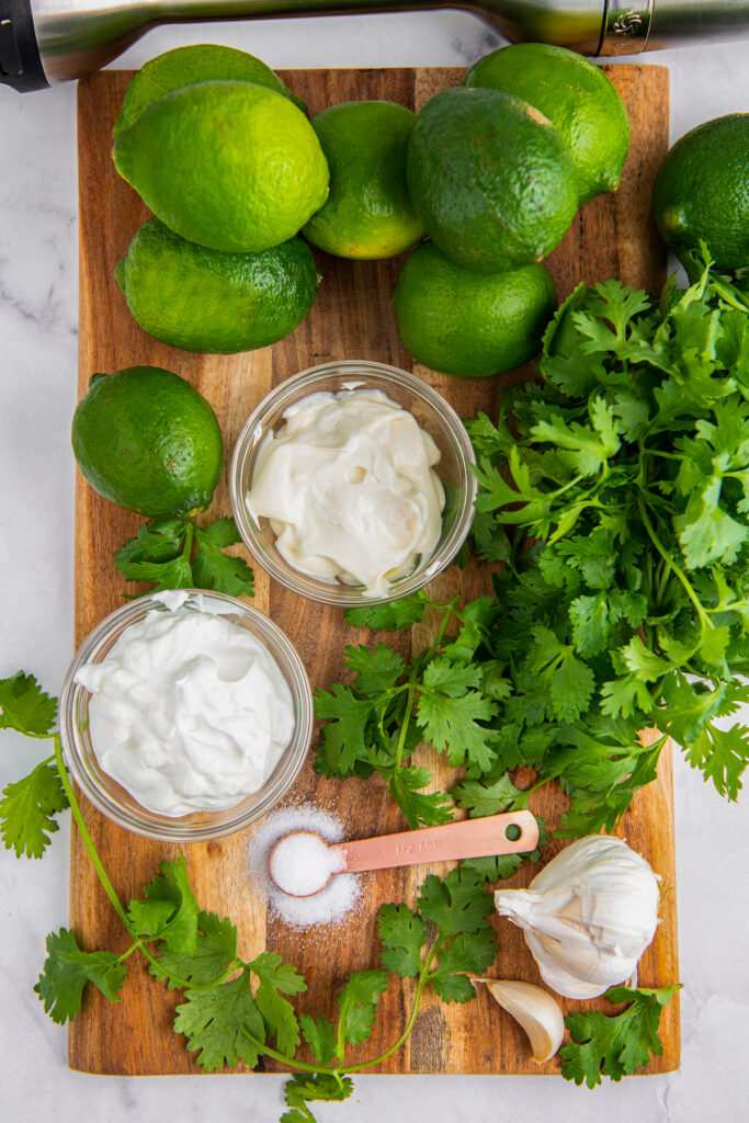 A wooden cutting board with limes, cilantro, garlic, salt, sour cream and mayonnaise 