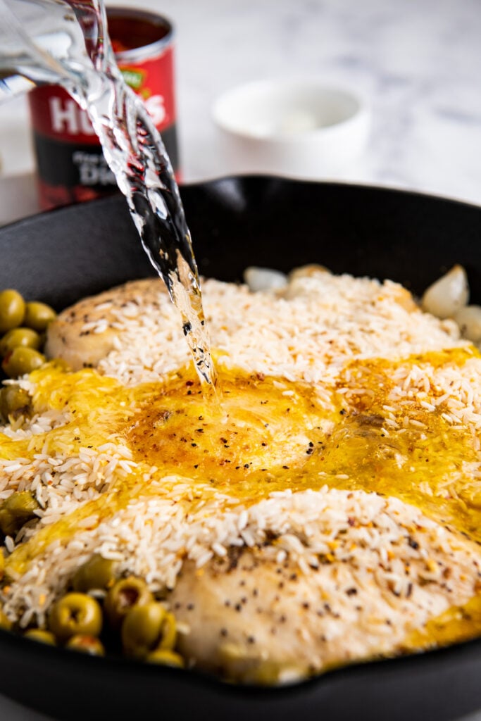 Water being poured over rice, chicken and olives and onions in a cast iron skillet