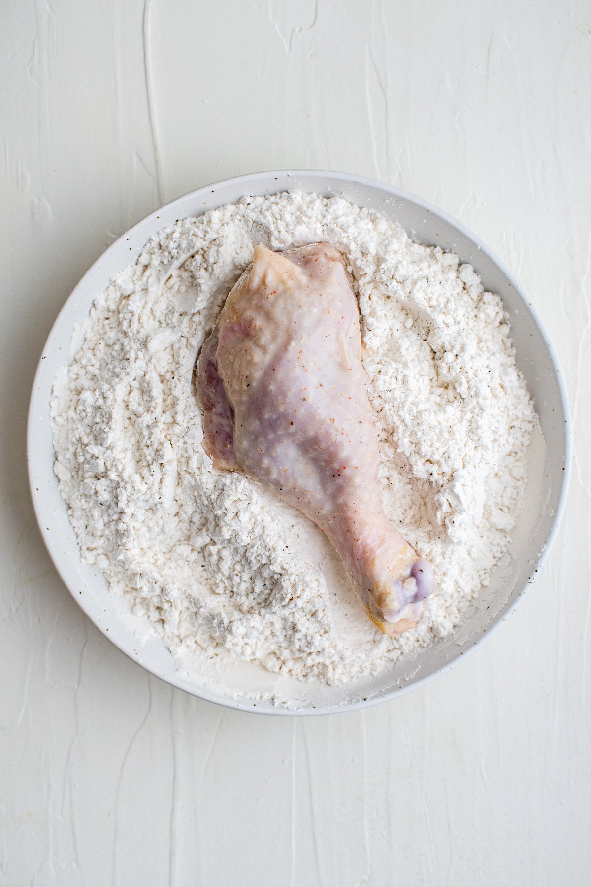 A chicken leg is placed in a white bowl filled with flour. 