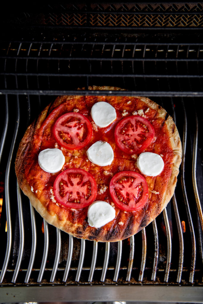 A pizza on a gas grill with sauce, sliced tomatoes and mozzarella cheese