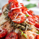 Side angle view of a chicken breast topped with melted cheese, pesto and roasted tomatoes.