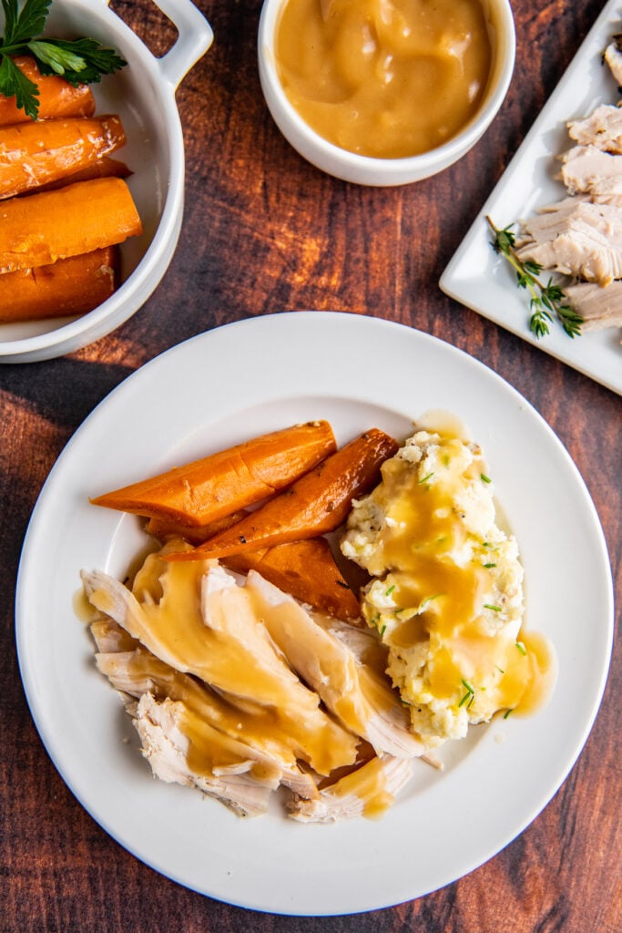 Overhead image of turkey breast sliced into pieces with gravy on top with carrots and mashed potatoes on a white plate.