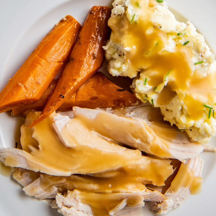 Overhead image of crockpot turkey breast sliced into pieces with gravy on top with carrots and mashed potatoes on a white plate.