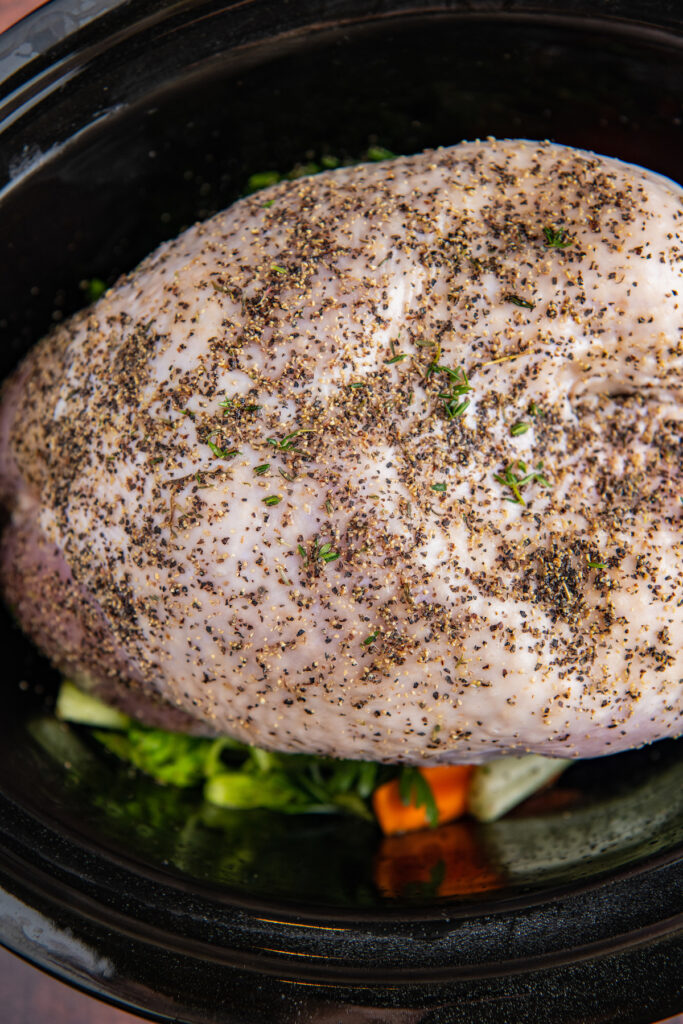 A turkey breast in a crockpot with herbs and carrots below it.