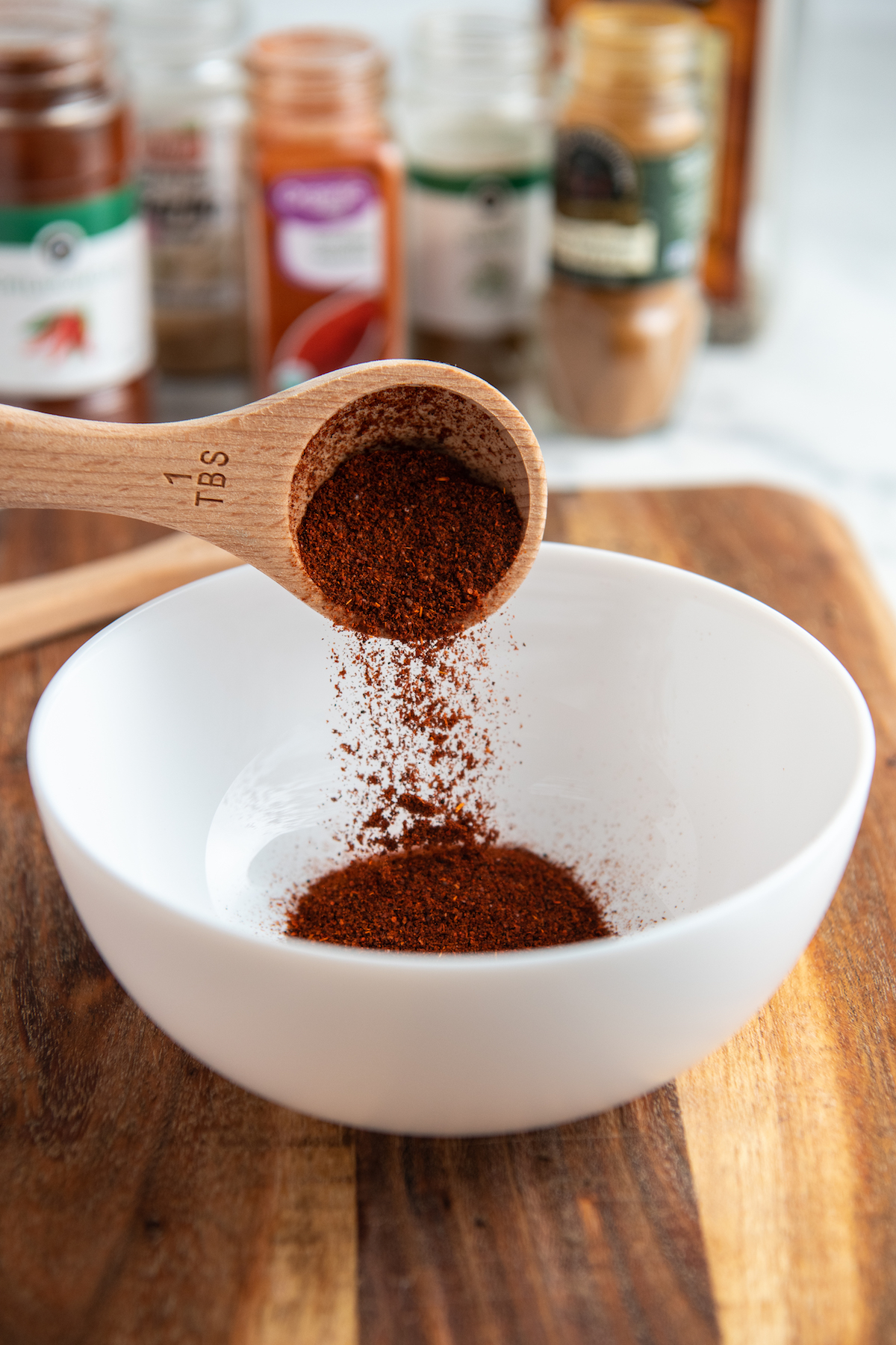 chili powder being poured into a white bowl with a wooden spoon.