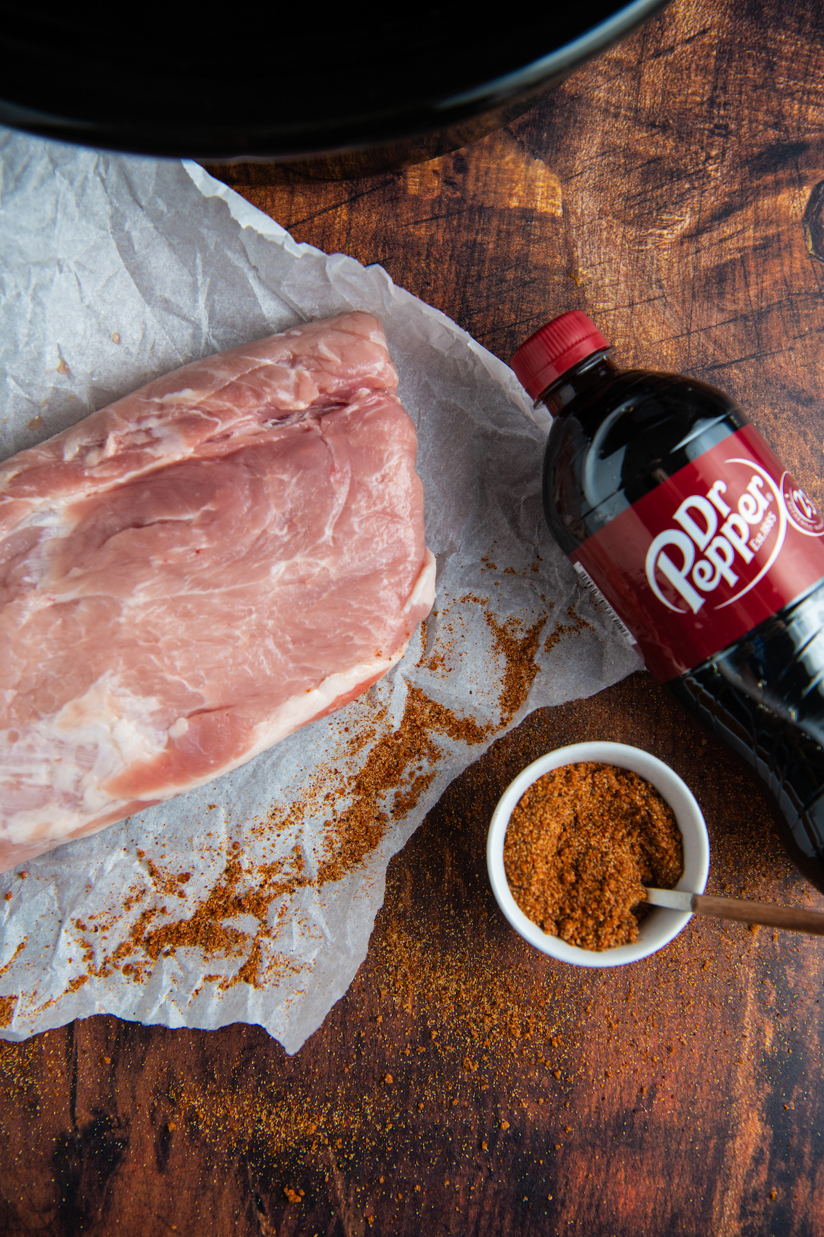 Overhead image of pork on parchment paper, a bottle of Dr Pepper and a jar with seasonings in it.