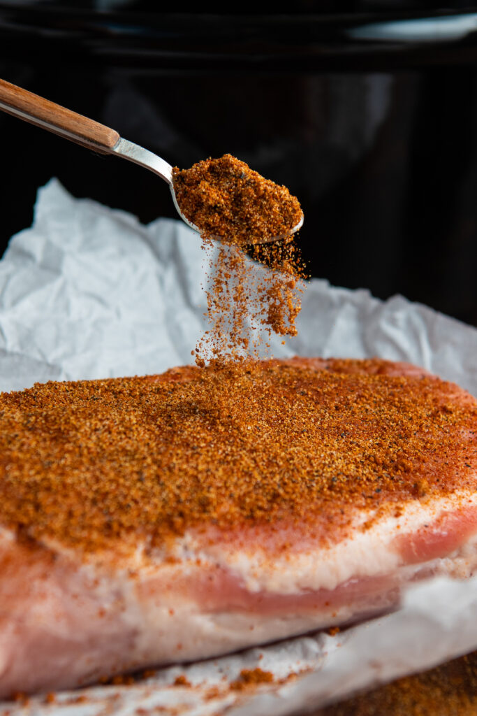 A piece of pork on parchment paper being sprinkled with seasonings.
