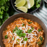 A bowl of mexican white chicken chili with spoons and lime wedges with a crockpot in the background.