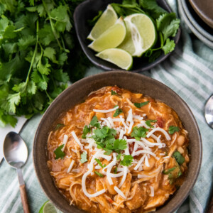 Mexican white chicken chili in a brown bowl with cheese and cilantro on top with lime wedges and a spoon.