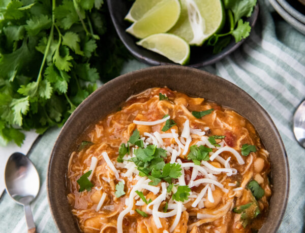 Mexican white chicken chili in a brown bowl with cheese and cilantro on top with lime wedges and a spoon.