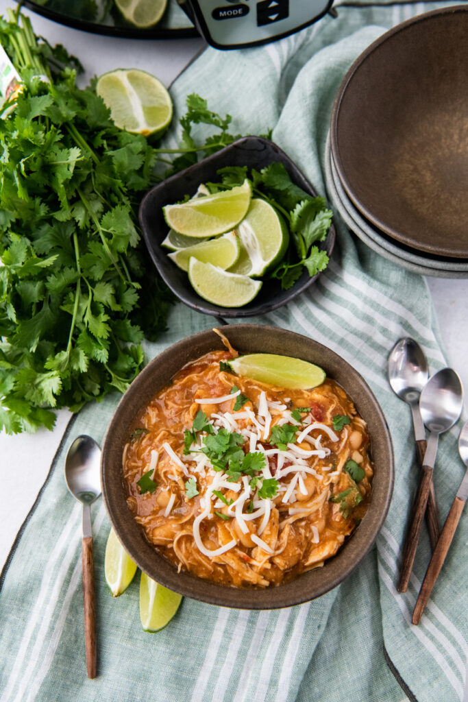 A bowl of mexican white chicken chili with spoons and lime wedges with a crockpot in the background.