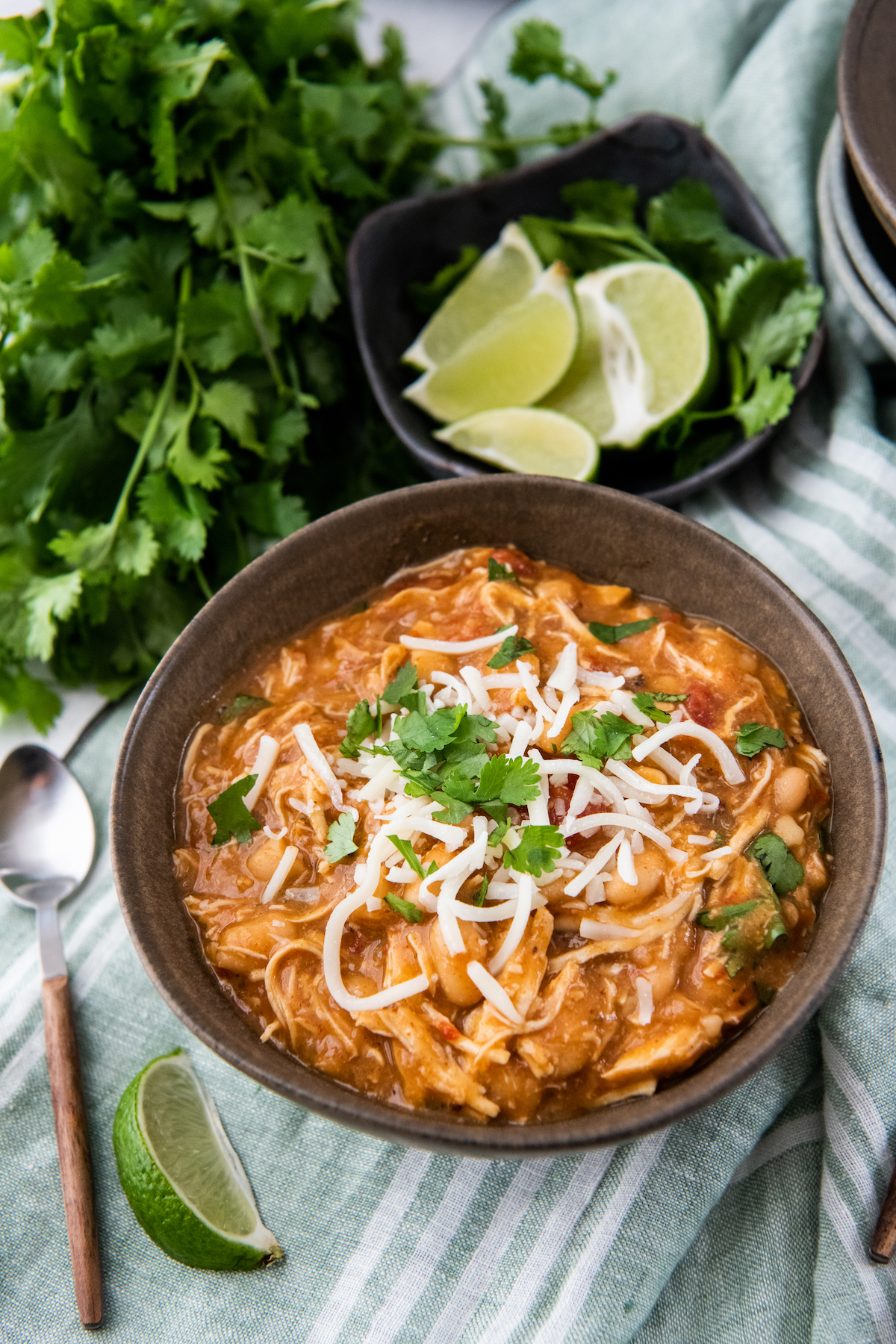 A bowl of crockpot white chicken chili in a bowl with cheese and cilantro on top with a spoon and cilantro in background.