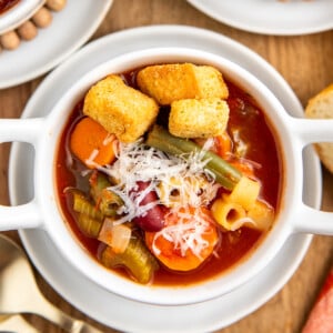 Up close image of minestrone soup in a white bowl with parmesan cheese and croutons on top.
